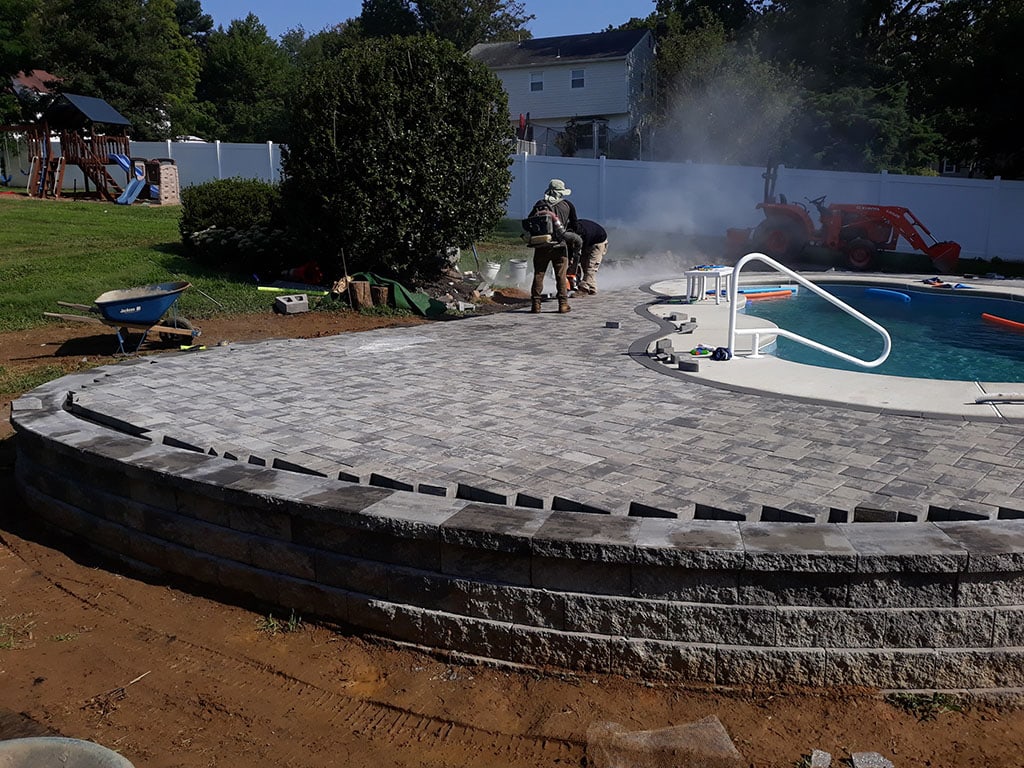 Workers are installing gray pavers as part of backyard hardscape design around a swimming pool, with construction equipment and a wheelbarrow nearby. Smoke or dust rises while they work, and a playground and house can be seen in the background.