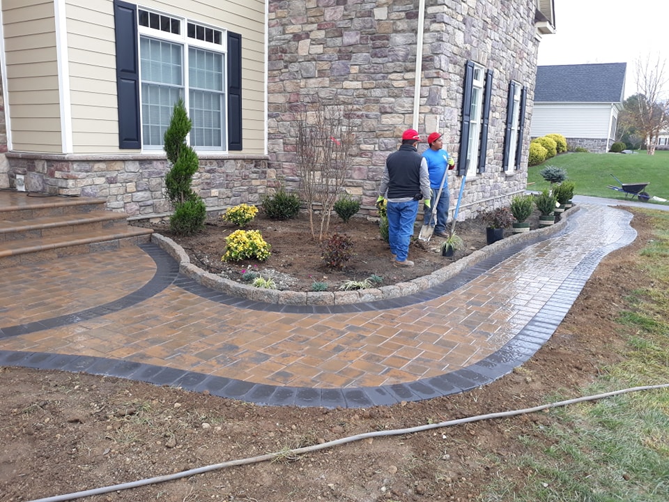 Three workers in red hats are landscaping beside a house with stone siding, installing a curved brick walkway as part of thoughtful hardscape design and planting shrubs and small trees along the edge.