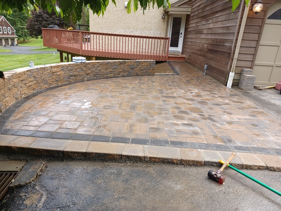A newly constructed patio with tan and dark gray pavers, bordered by a curved stone masonry retaining wall beside a brown wood-sided house and a red-railed porch. A mallet and green garden hose lie on the asphalt nearby.