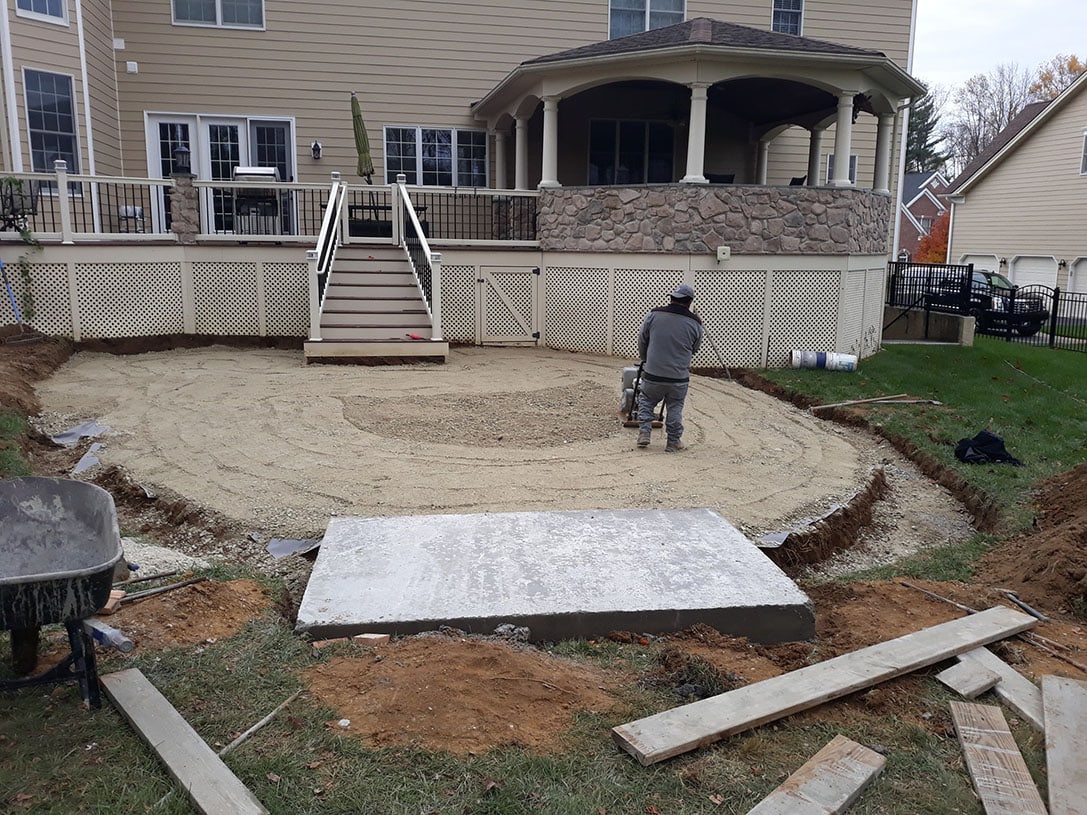 A person operates a compactor on a circular area of leveled dirt in a backyard, preparing the ground for hardscape design near a house with a raised deck and stairs. Construction materials are scattered nearby.