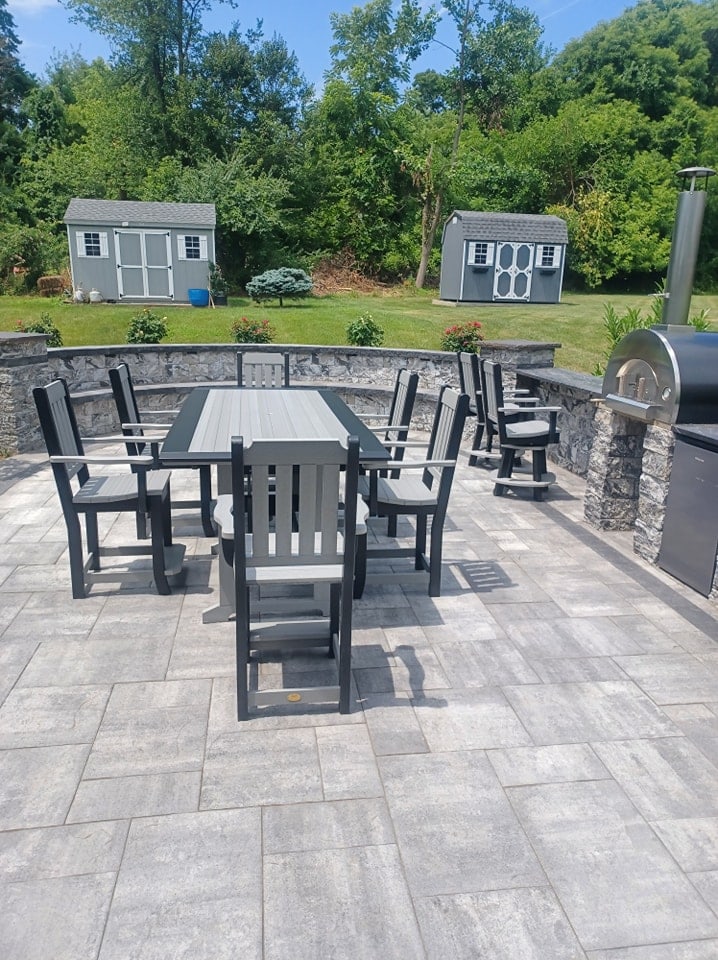 Outdoor patio with a rectangular dining table and eight chairs on gray stone tiles. The landscaping features two gray storage sheds in the background, set against lush green trees and shrubs.