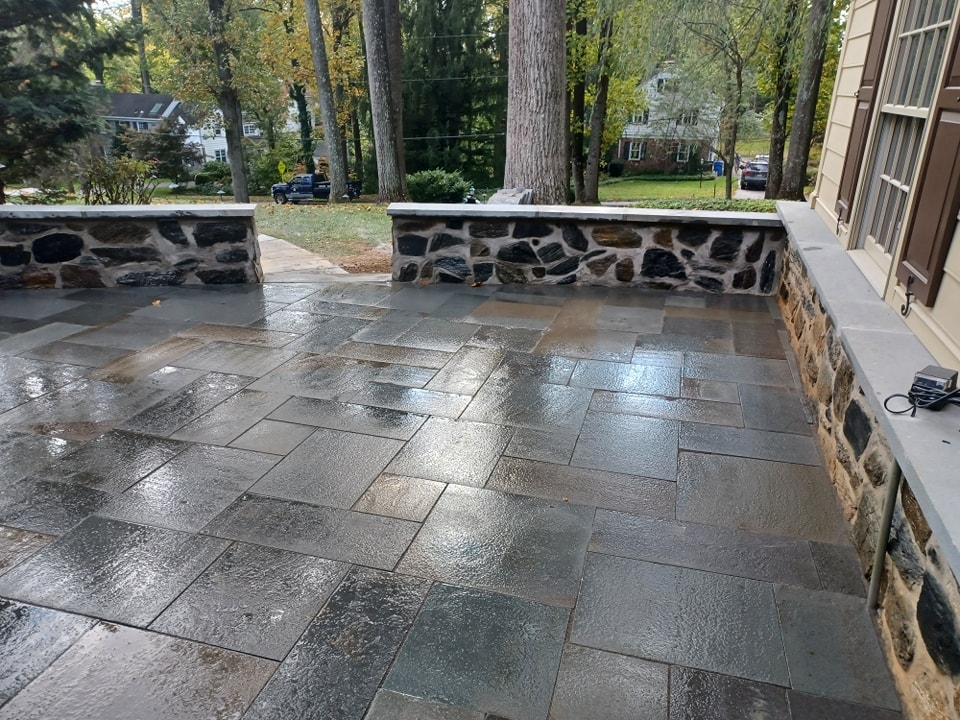 A wet stone patio with dark rectangular tiles showcases elegant masonry, bordered by low stone walls. Tall trees and residential houses are visible in the background, enhancing the landscaping appeal.