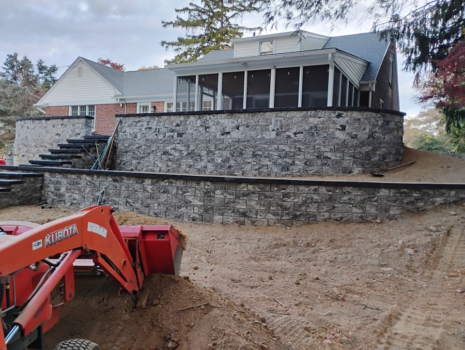 A newly constructed masonry retaining wall with stairs leads up to a house with a large screened porch. A red Kubota tractor is parked on the dirt slope in the foreground, highlighting the fresh landscaping. Trees are in the background.