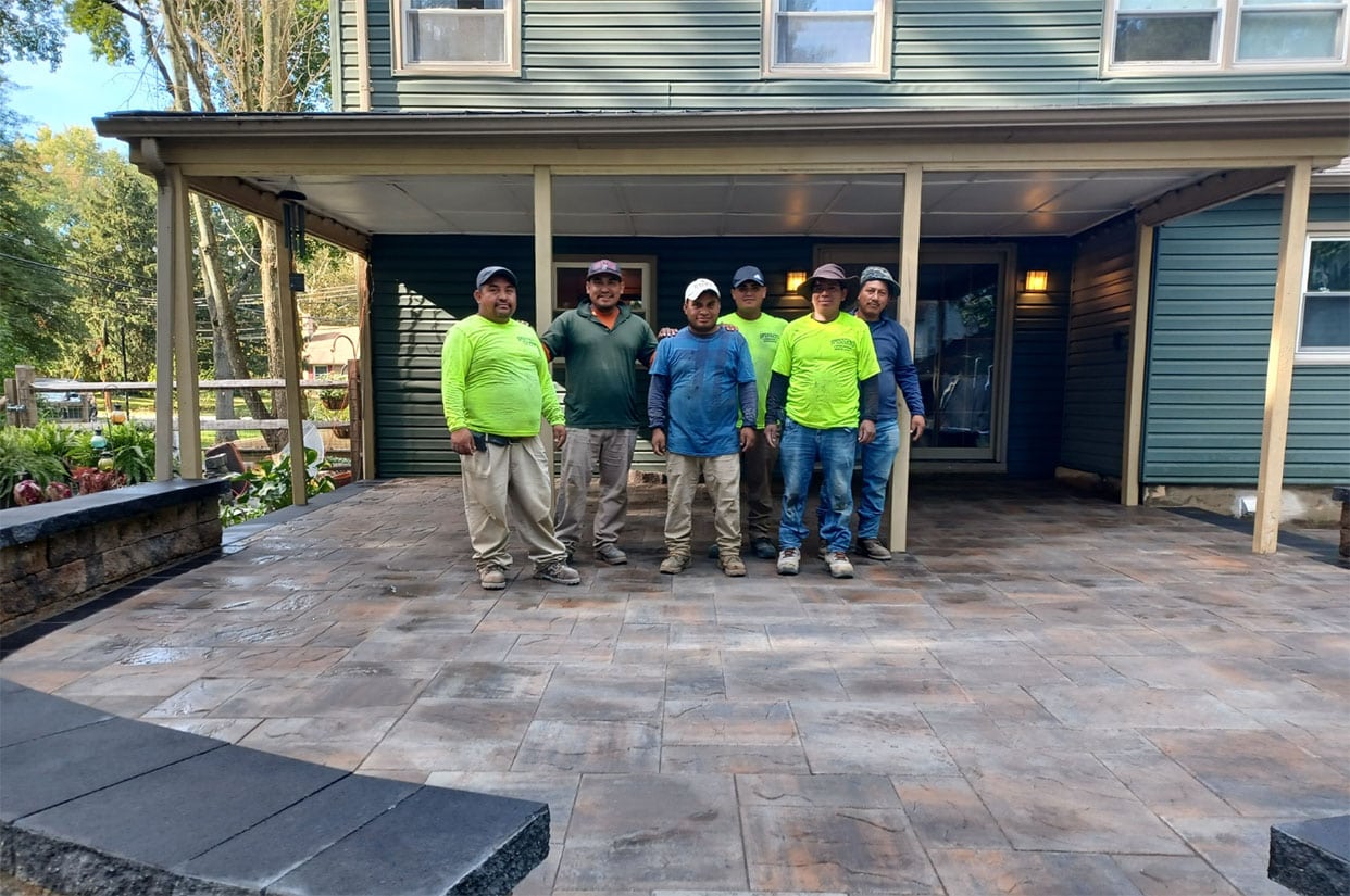 Six construction workers stand together on a newly completed stone patio, showcasing their masonry and hardscape design skills, in front of a green house with tan trim and a covered porch. They wear work clothes, some with hats or bright safety shirts.
