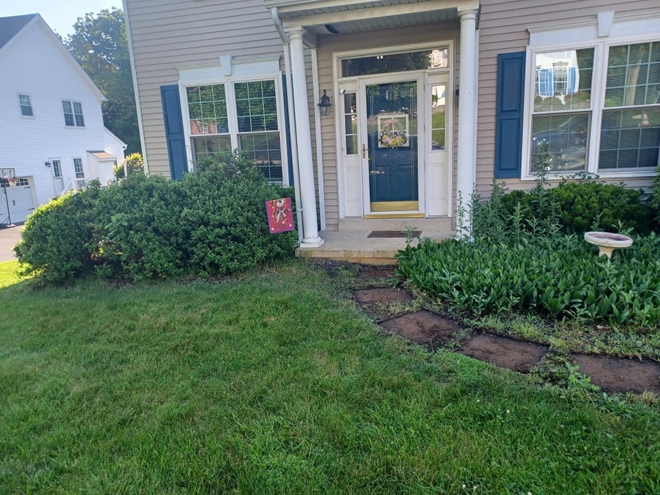 A front yard garden featuring lush landscaping with green bushes, a small stone path leading to a porch with white pillars, and a yellow-trimmed door. A red garden sign and birdbath add charm to the inviting hardscape design.