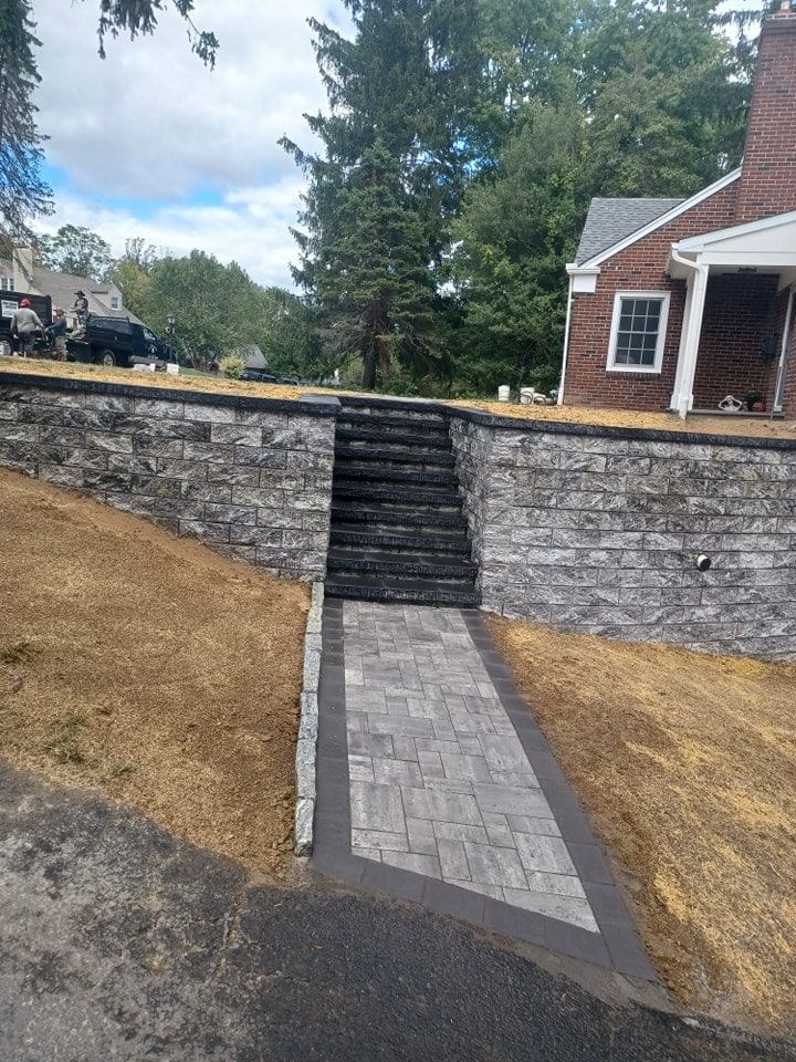 A stone walkway with dark borders showcases expert masonry as it leads up bordered steps, flanked by stone retaining walls, to a grassy yard in front of a brick house with a porch. Trees and other homes can be seen in the background.