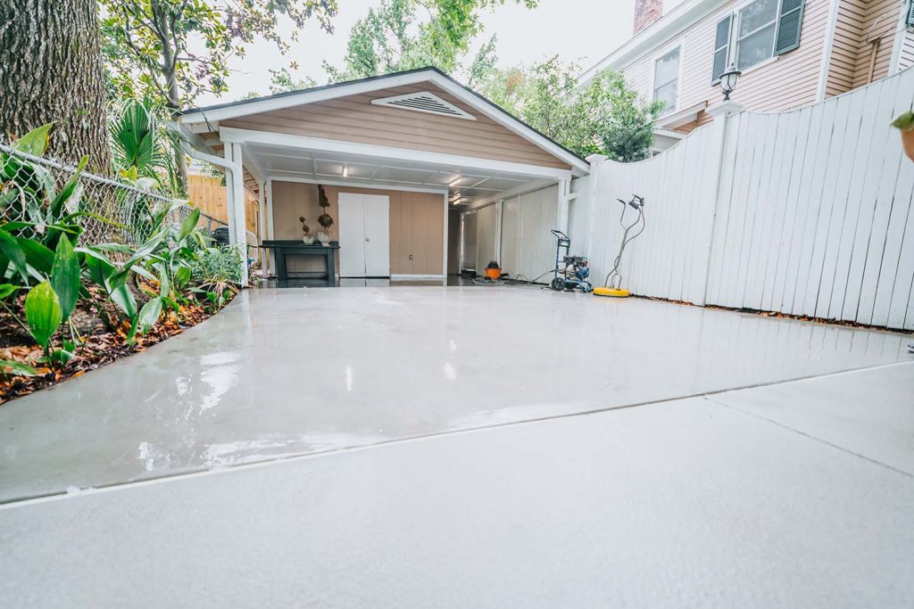 A freshly poured and smoothed concrete driveway leads to a tan garage with white trim, surrounded by a white fence, lush landscaping, green plants, and garden tools along the side.