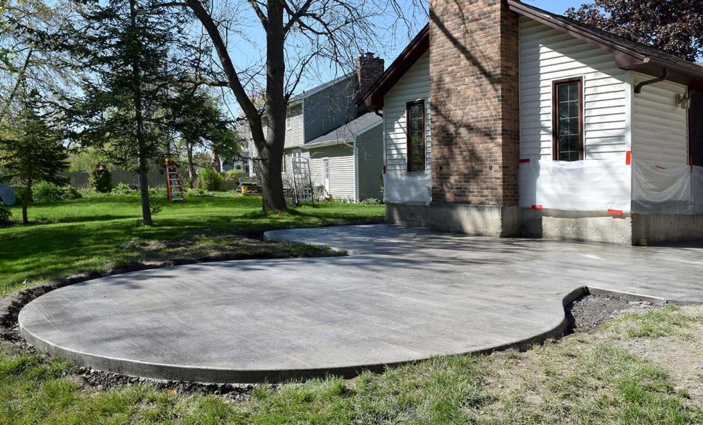 A freshly poured concrete patio with curved edges extends from a house with white siding and brick accents, showcasing thoughtful landscape design surrounded by grass, trees, and a neighboring home.