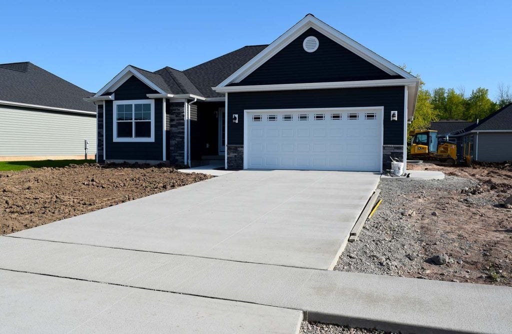 A newly constructed house with dark siding and a white garage door features a large, fresh concrete driveway. The yard, still bare soil, is ready for future landscaping and hardscape design to complete the home's exterior.
