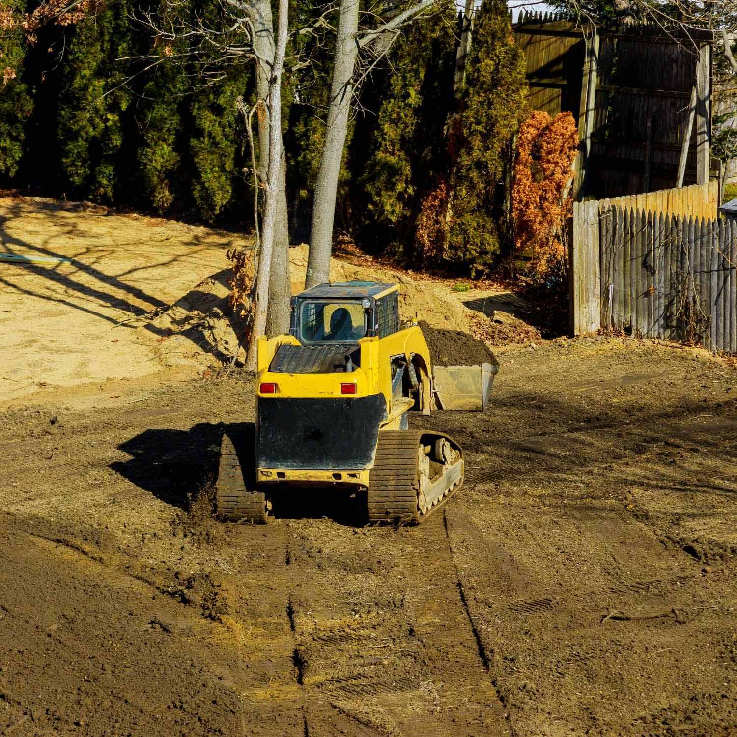 A yellow and black bulldozer moves earth in a yard, creating tracks in the soil near trees and a wooden fence on a sunny day, preparing the site for landscape design.