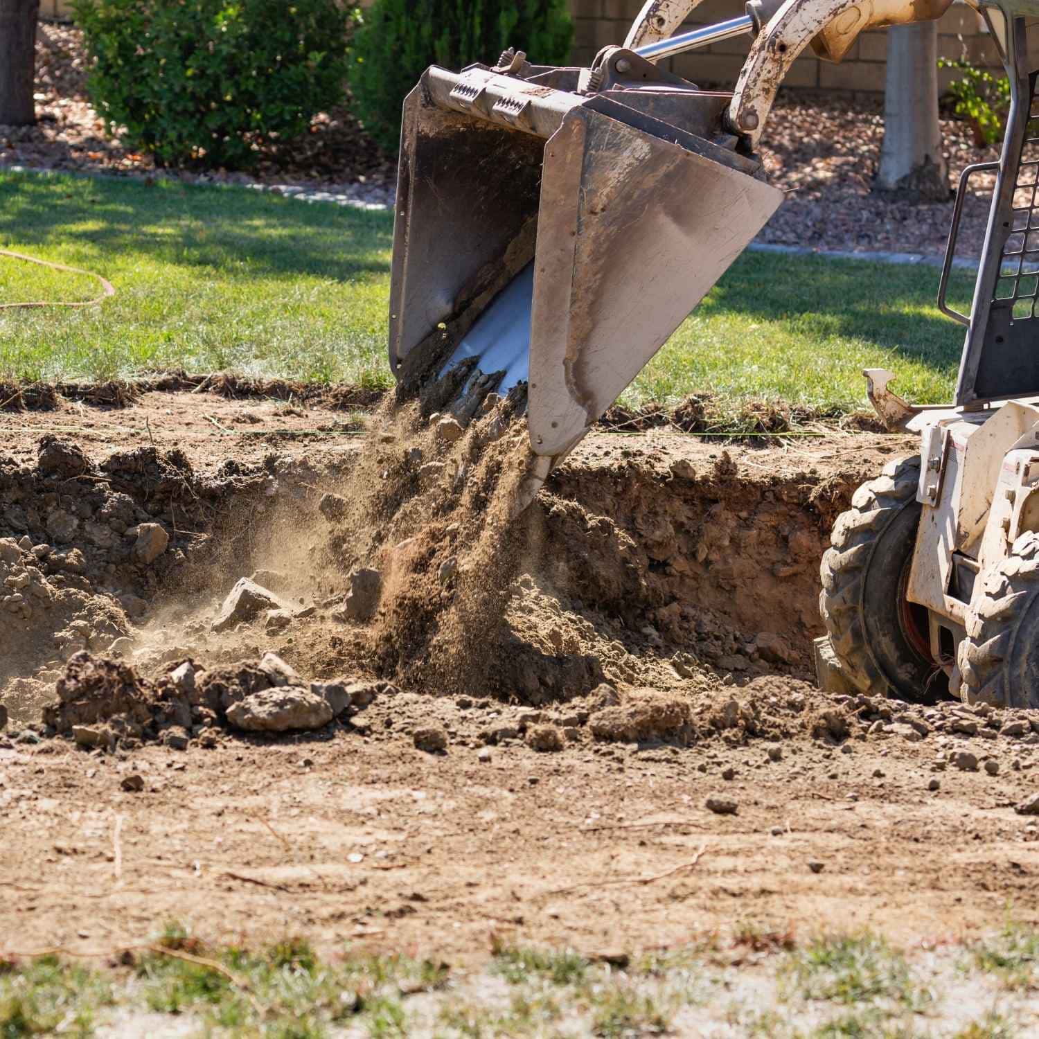 A construction vehicle dumps dirt into a dug-out area in a yard, preparing the site for upcoming landscaping, with grass and trees visible in the background.