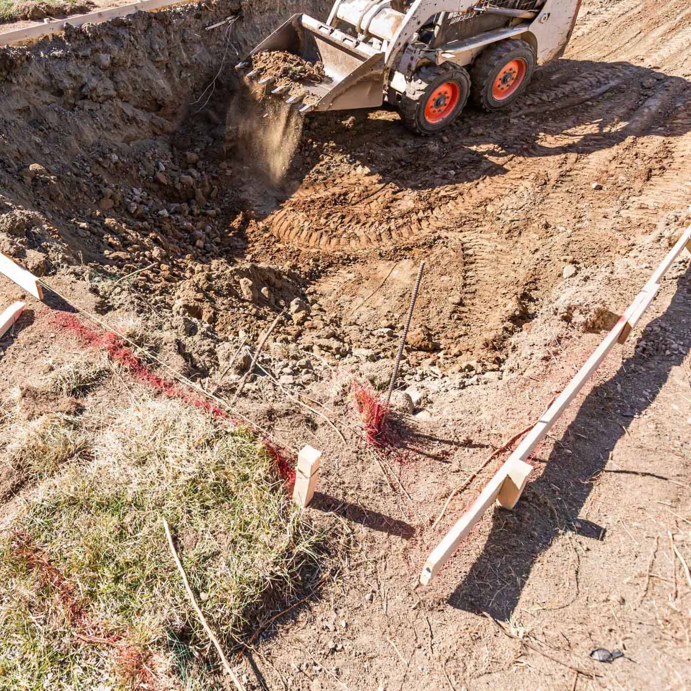 A bulldozer moves dirt in a construction site, creating a large hole in the ground marked by wooden stakes and red string—preparing the area for future hardscape design and landscaping.