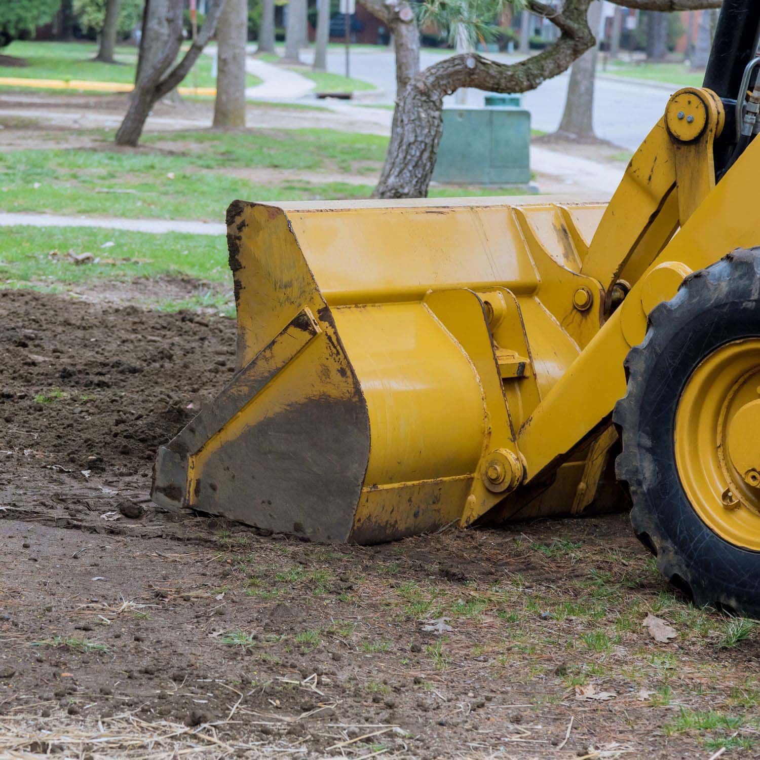 Close-up of a yellow bulldozer's front bucket digging and moving soil in a park-like area, preparing the ground for landscape design, with grass, trees, and a sidewalk visible in the background.