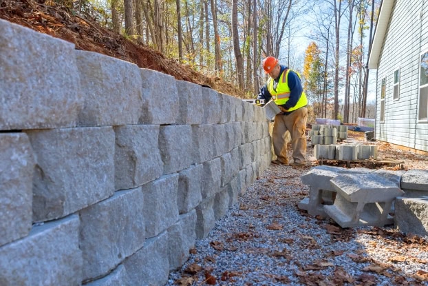 A construction worker in a safety vest and helmet measures a newly built stone retaining wall, showcasing expert hardscape design next to a house. Tools and concrete blocks rest on the gravel path, while trees enhance the landscaping in the background.