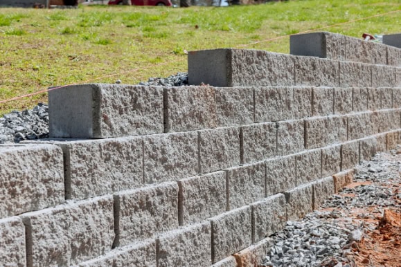 A close-up view of a newly constructed masonry retaining wall made of stacked concrete blocks, with gravel at the base and grass on the slope above, showcases expert hardscape design.