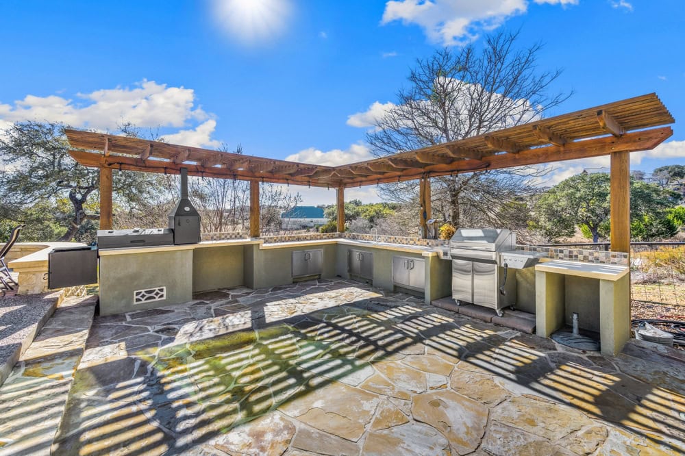 Outdoor kitchen with a wooden pergola, stone floor, built-in grills, sink, and counter space showcases thoughtful hardscape design in a sunny backyard. Shadows from the pergola create patterns on the floor amid lush trees and a clear blue sky.