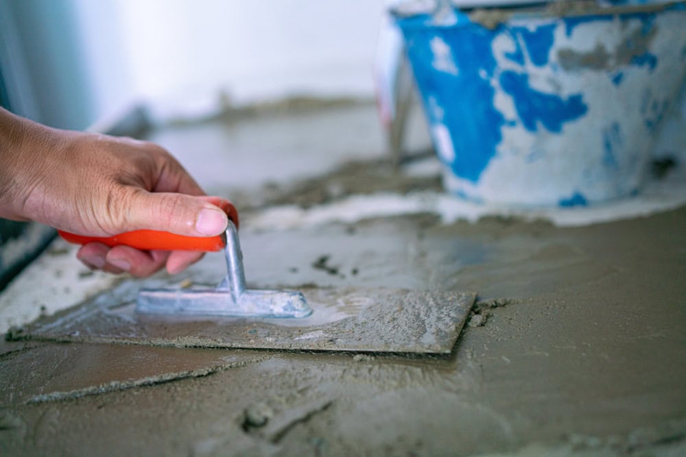 A close-up of a hand using a trowel to smooth wet cement, with a blue bucket in the background. The scene suggests hardscape design or home improvement as part of a larger landscaping project.
