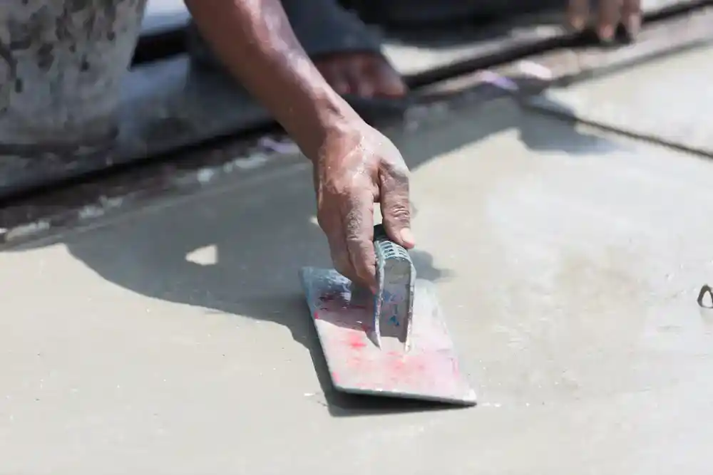 A close-up of a person’s hand using a metal trowel to smooth wet concrete on a flat surface—an essential step in masonry and hardscape design—with another hand visible in the background.