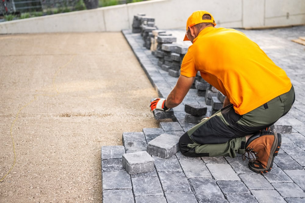A worker in an orange shirt, cap, gloves, and boots kneels on freshly laid gray paving stones, skillfully arranging bricks as part of a hardscape design to construct a pathway or patio in an outdoor landscaping project.
