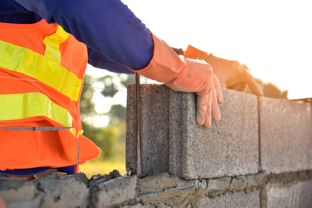 A construction worker wearing an orange safety vest and gloves places a concrete block onto a wall under construction, showcasing expert masonry skills with sunlight in the background.