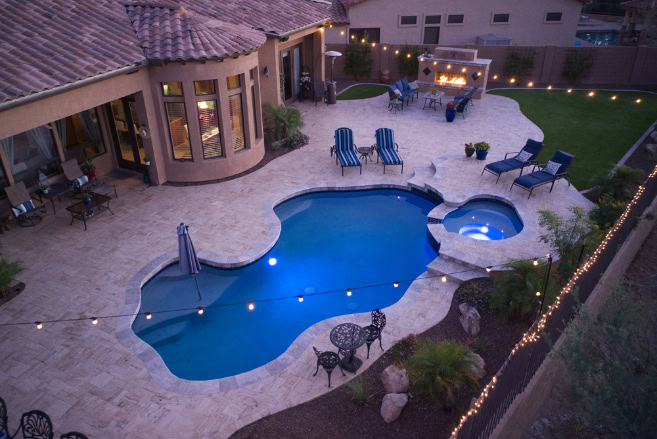 Aerial view of a backyard with a curvy pool and spa, lounge chairs, string lights, outdoor dining area, barbecue grill, meticulous landscaping, green lawn, and a tan tiled patio beside a modern house at dusk.