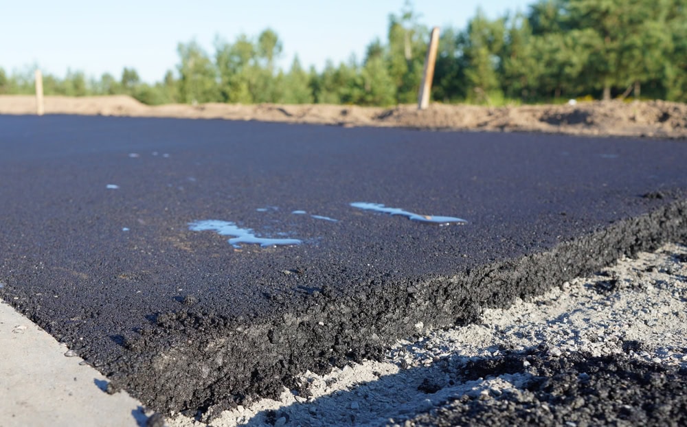 Close-up of freshly laid asphalt pavement, with small puddles of water on the surface. The background features a blurred construction site with soil, trees, and clear blue sky—perfect inspiration for hardscape design and landscaping projects.