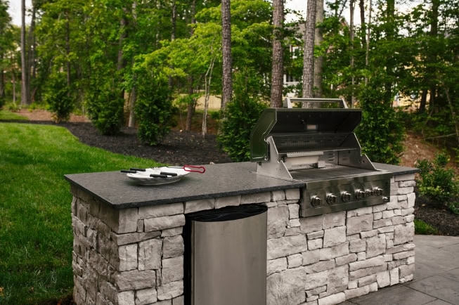 An outdoor stone grill station showcasing expert masonry and a stainless steel grill, trash bin, and grilling utensils on the countertop, set in a green backyard surrounded by trees—a perfect addition to any landscape design.