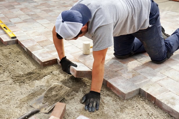 A person wearing gloves and a cap is kneeling on the ground, carefully laying rectangular bricks to create a paved surface—a clear display of skilled masonry. Tools and sand are visible nearby.