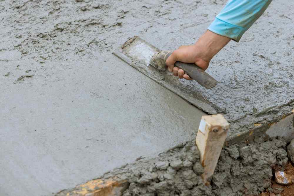 A person smooths wet cement with a trowel, wearing a light blue long-sleeve shirt—capturing the careful attention to detail in hardscape design as the hand and tool work on a freshly poured concrete surface.