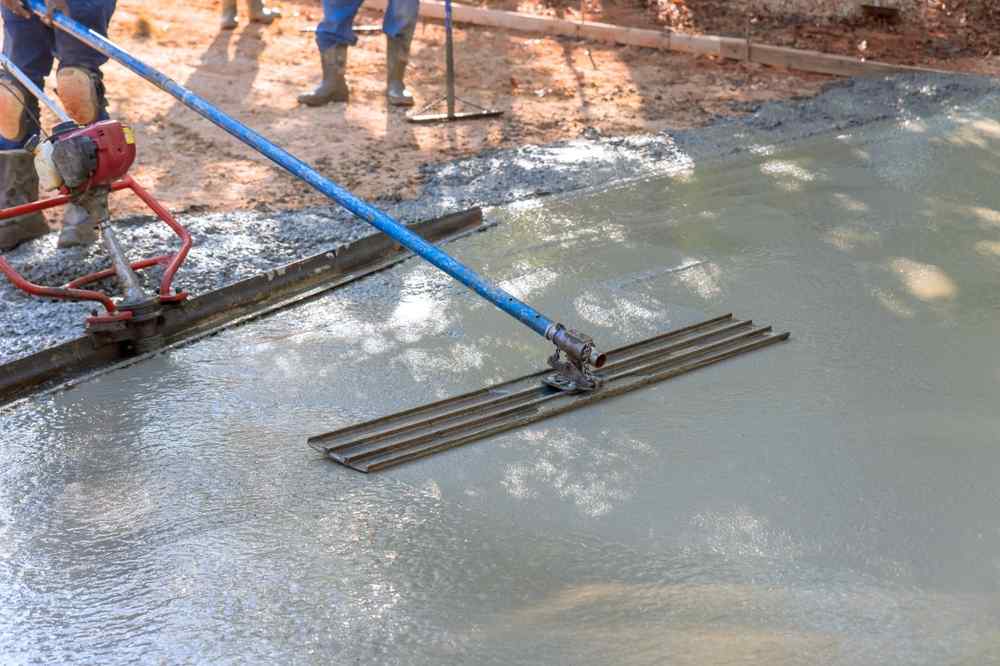 A person uses a long-handled tool to smooth and level freshly poured concrete on a construction site, showcasing essential masonry skills, while others stand nearby on the gravel.