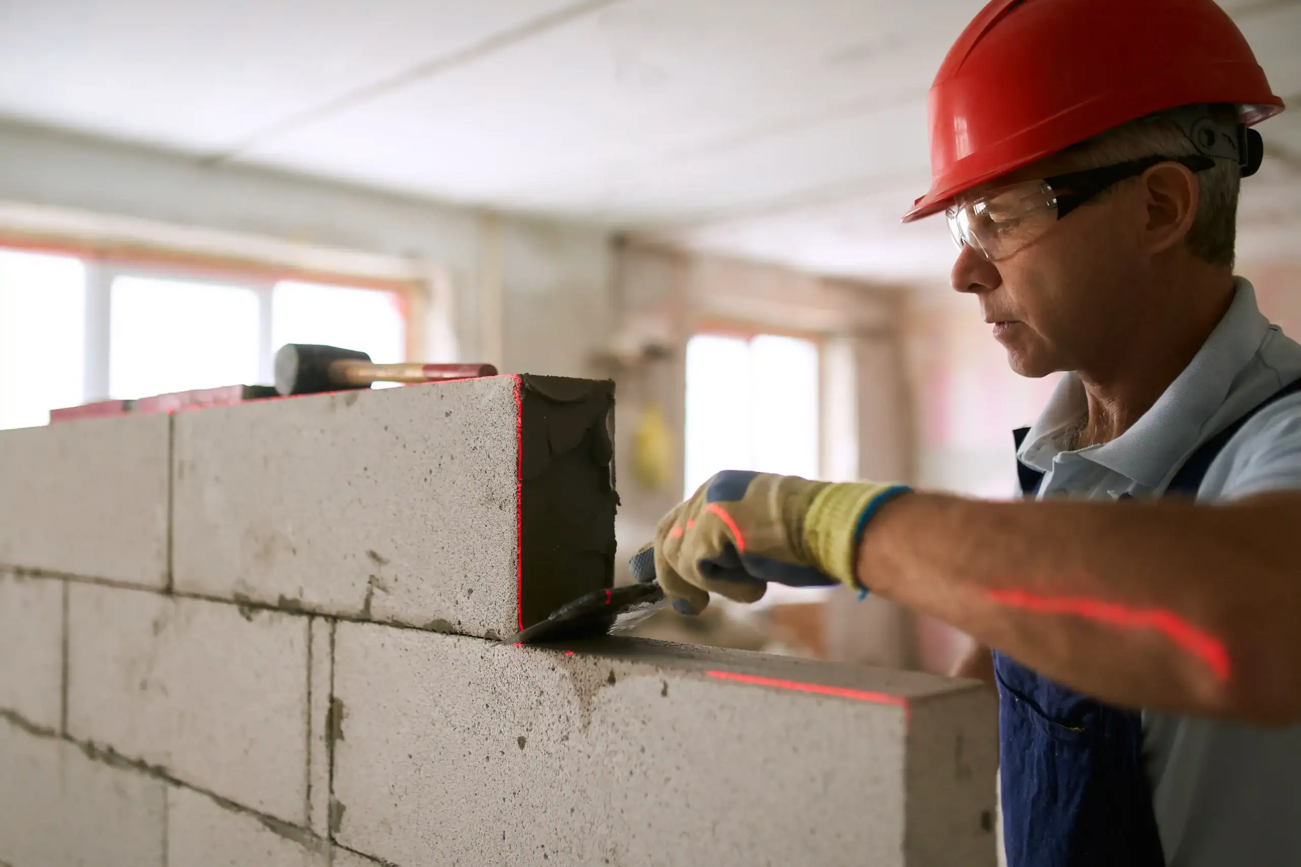 A construction worker wearing a red hard hat and safety glasses carefully lays concrete blocks with mortar, showcasing skilled masonry as he uses a trowel to smooth the joints while building a wall inside a well-lit building under construction.