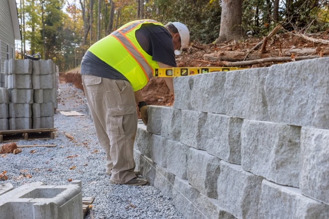 A construction worker in a neon safety vest and cap uses a level to check the alignment of large gray stone blocks while building a masonry retaining wall outdoors. Trees and stacked blocks create a natural landscaping backdrop.