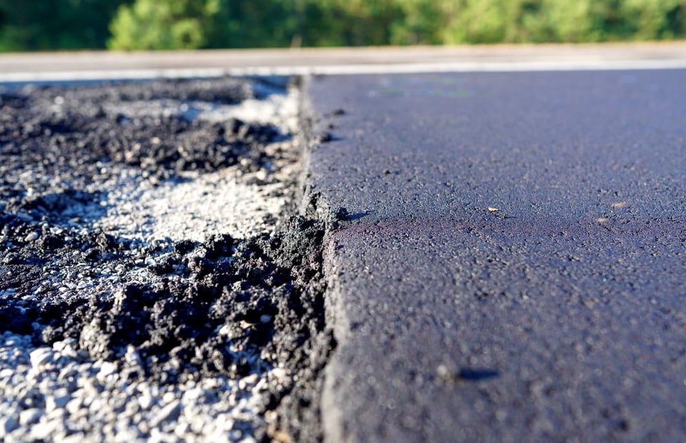 Close-up view of a newly paved asphalt road with a sharp edge, contrasting with older, rougher asphalt; blurred greenery suggests thoughtful landscape design in the background.