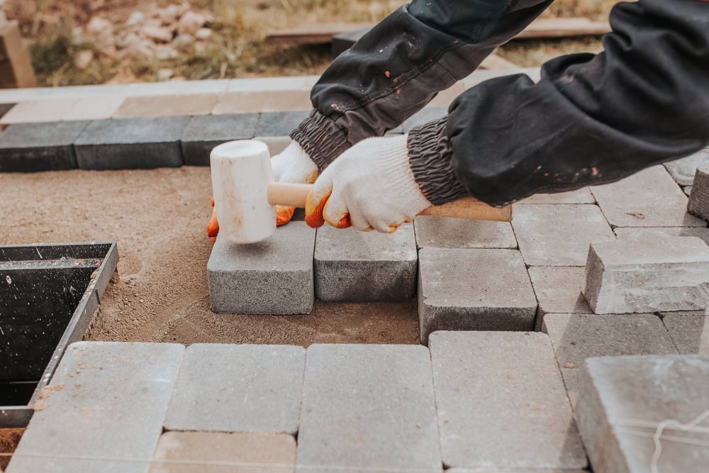 A person wearing gloves uses a rubber mallet to adjust grey paving stones while laying a pathway outdoors, showcasing skilled masonry and thoughtful hardscape design.