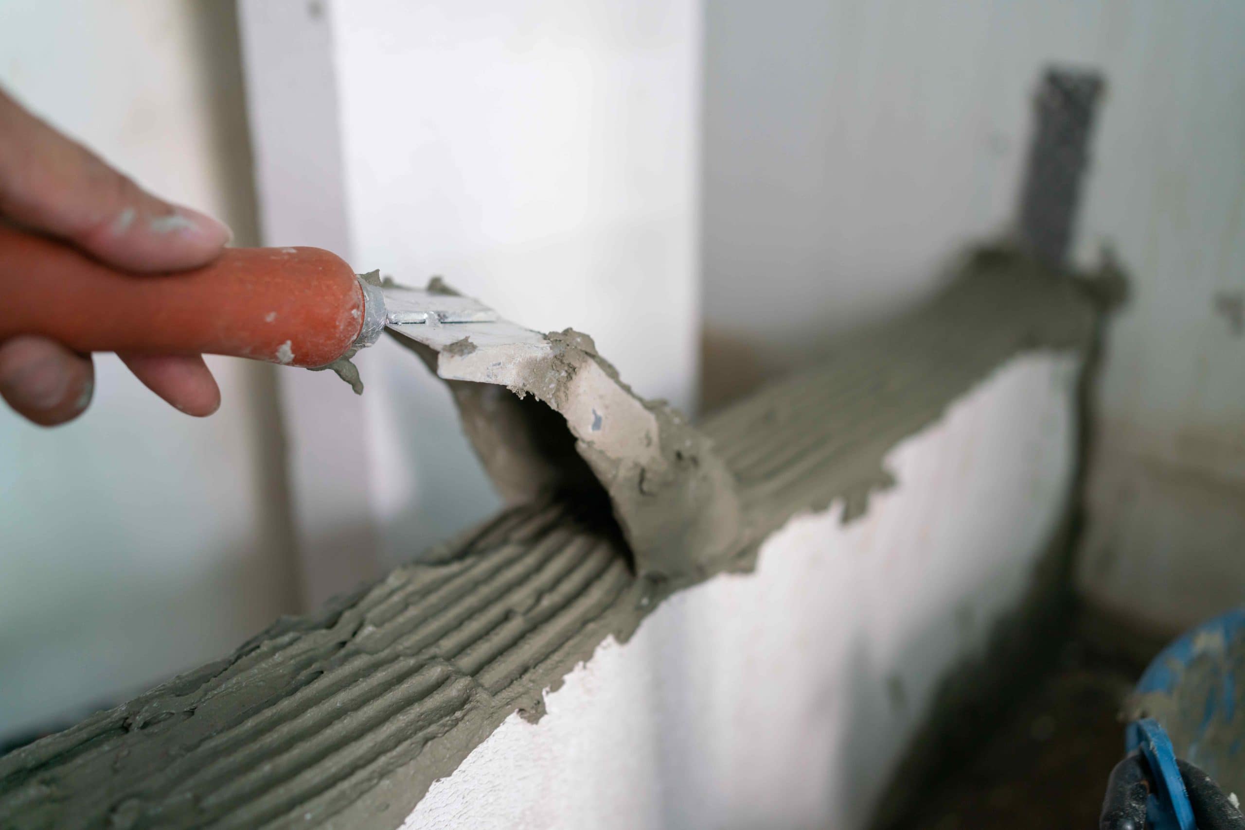 A hand holding a trowel spreads wet cement on top of a white brick wall, smoothing the surface for masonry work in landscaping or hardscape design.