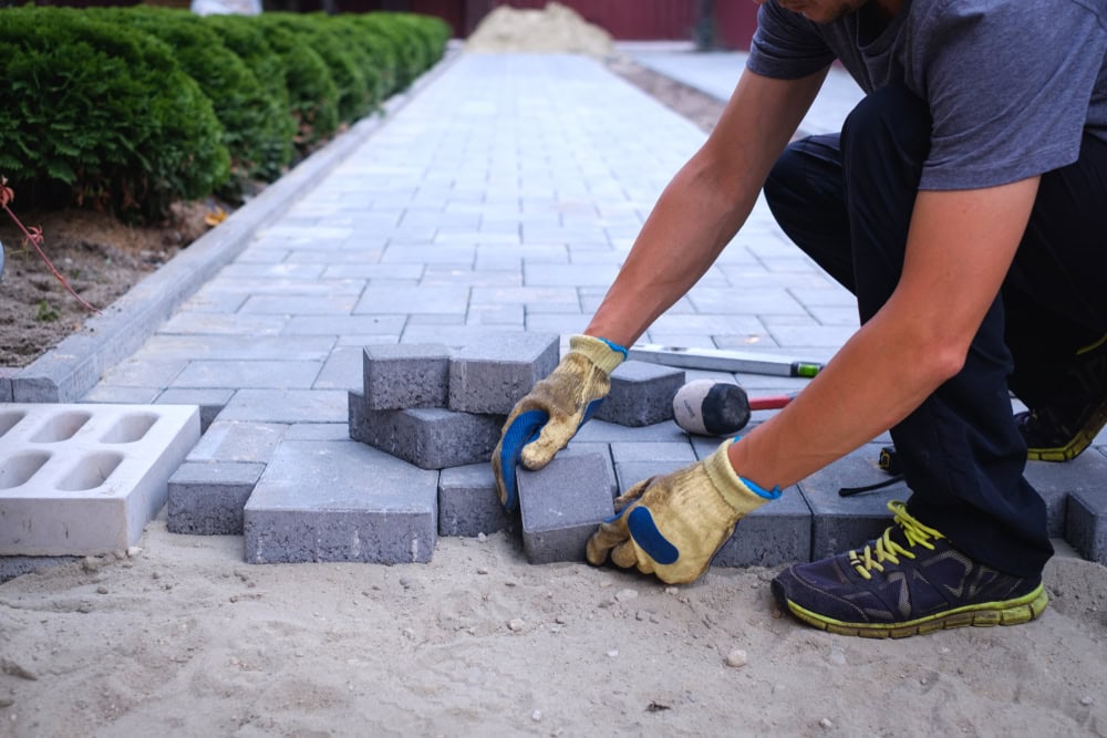A person wearing gloves and sneakers places rectangular gray paving stones onto a sandy surface, demonstrating expert masonry as they construct a patterned walkway with bushes lining one side. Tools are scattered nearby.