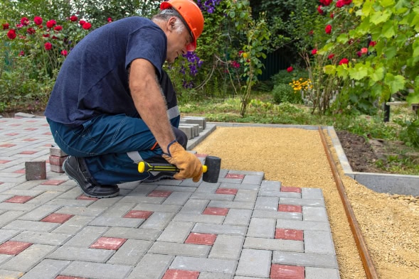 A worker wearing a hard hat is kneeling and installing gray and red paving stones in a garden area, using a rubber mallet and level, showcasing expert hardscape design with flowers and plants in the background.