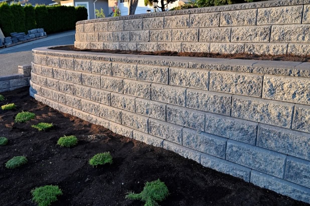 Tiered retaining wall made of textured gray stone blocks, supporting a landscaped garden with green plants and dark soil; road and hedges in the background.
