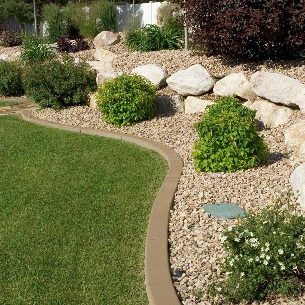 A landscaped garden featuring neatly edged green grass, curved concrete borders, and large decorative rocks set among gravel highlights expert hardscape design. Small bushes, white flowers, shrubs, and plants complete the tranquil setting.