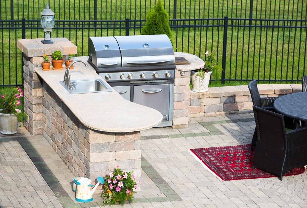 Outdoor kitchen with a stone countertop, built-in sink, stainless steel grill, potted plants, and a patio dining set on a rug—an inviting masonry feature that elevates the landscaping next to a black metal fence and green lawn.
