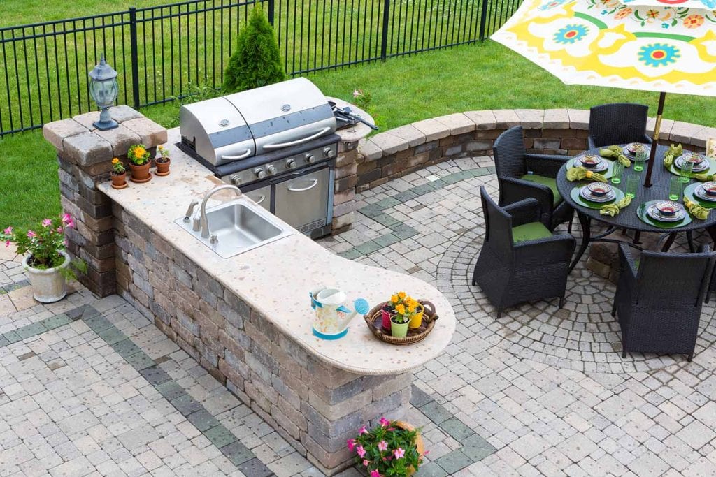 Outdoor patio features masonry elements like a built-in stone grill and sink, surrounded by potted flowers. A round table for six sits under a colorful umbrella, all set within lush landscaping and bordered by a black fence.