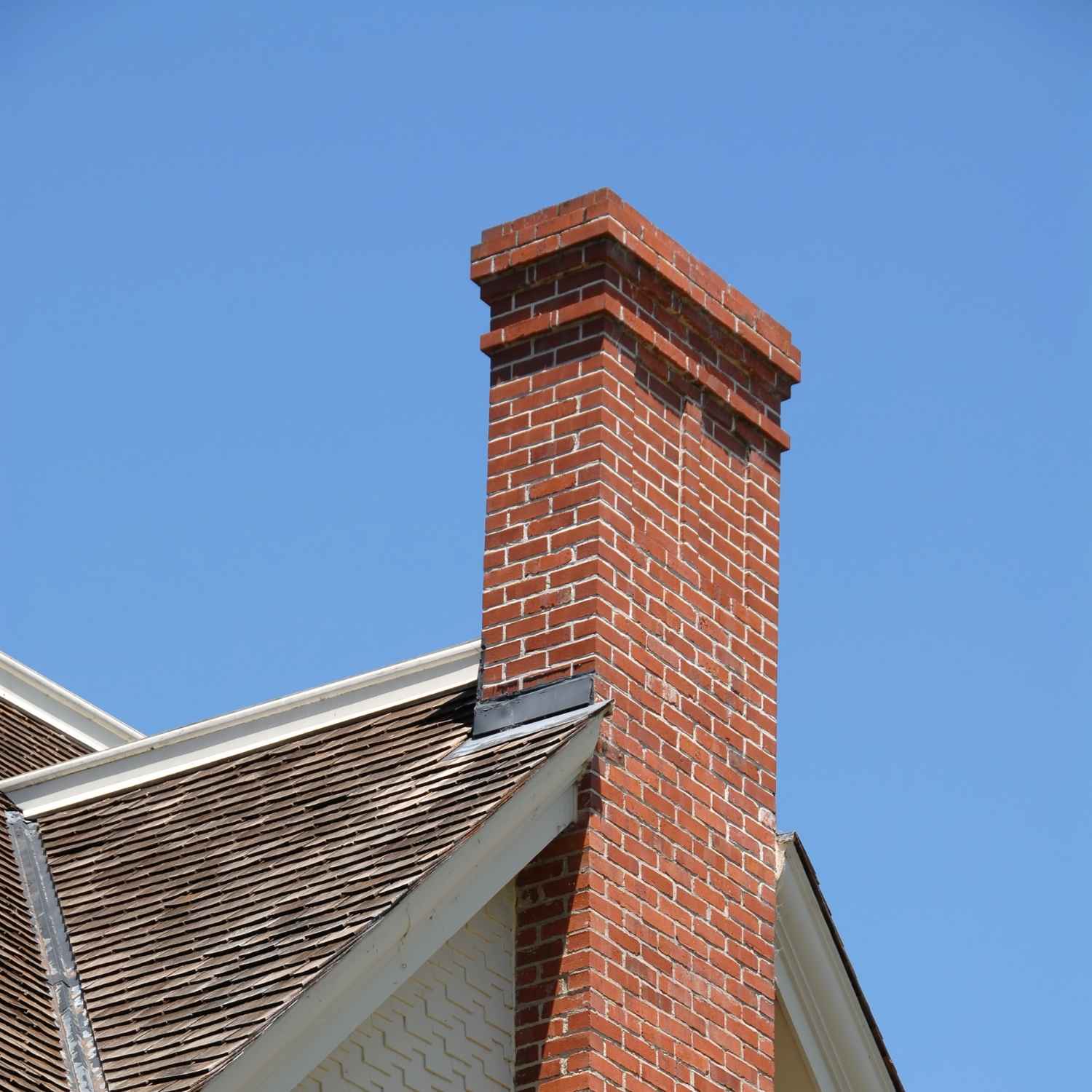 A red brick masonry chimney rises above a sloped roof with wooden shingles under a clear blue sky.