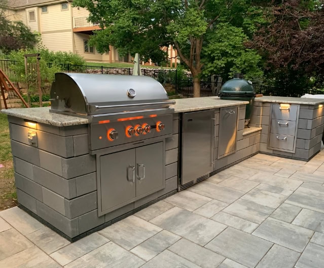 Outdoor kitchen with stainless steel appliances, stone countertop, and built-in lights features expert masonry and hardscape design on a stone patio, surrounded by trees and a fenced yard for seamless landscape design integration.