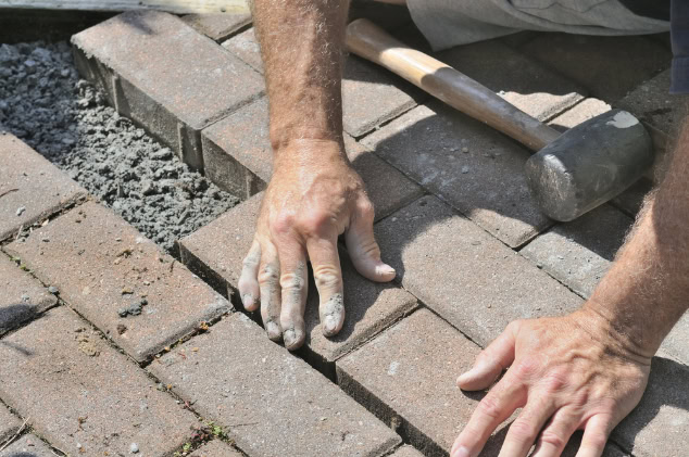 A person’s hands are adjusting brick pavers into place on the ground, with a rubber mallet nearby and sand or gravel in the gaps—a detailed look at hardscape design and patio construction in progress.