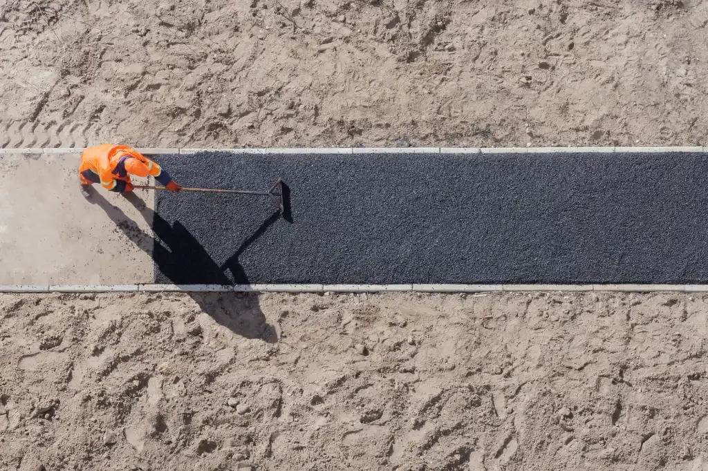 Top-down view of a construction worker in an orange uniform spreading fresh black asphalt on a narrow dirt-bordered pathway, casting a long shadow.
