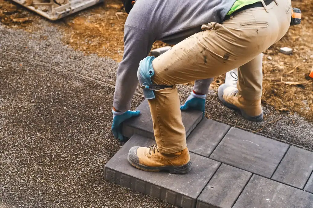 Construction worker wearing gloves, knee pads, and boots installing rectangular concrete pavers on a compacted gravel base.
