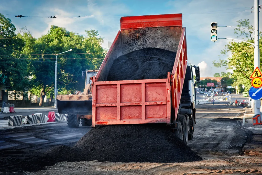 A red dump truck unloads asphalt as Paving Contractors Delaware County, PA work near trees and signs.